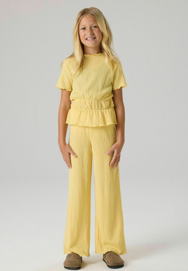 Jeune fille debout et souriante, portant un haut jaune texturé à manches courtes avec un détail volanté et un pantalon large assorti avec des chaussures marron.