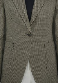 Gros plan sur un blazer à motif pied-de-poule avec un bouton noir unique et deux poches avant, porté sur un haut noir et un pantalon blanc.