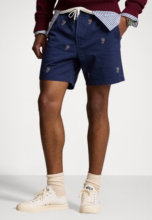 Mand iført marineblå shorts med små broderede logoer, en burgunderrød sweater over en stribet skjorte, cremefarvede sokker og hvide sneakers, stående.