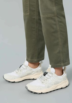 Weiße und cremefarbene Sneaker mit texturiertem Obermaterial, das mit floralem Lochmuster und grauer Gummisohle versehen ist. Kombiniert mit olivgrünen, umgeschlagenen Hosen.