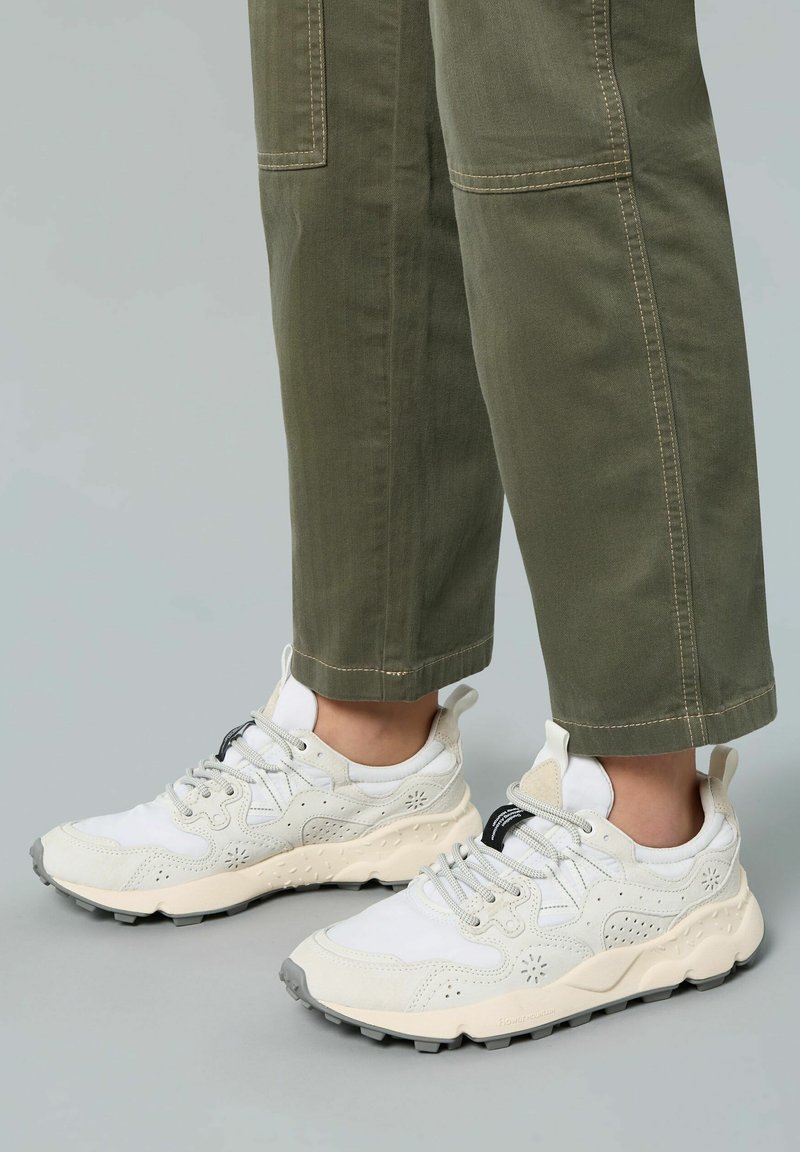 Sneakers bianchi e crema con una parte superiore testurizzata, caratterizzati da perforazioni floreali e suola in gomma grigia. Abbinati a pantaloni verde oliva con risvolto.