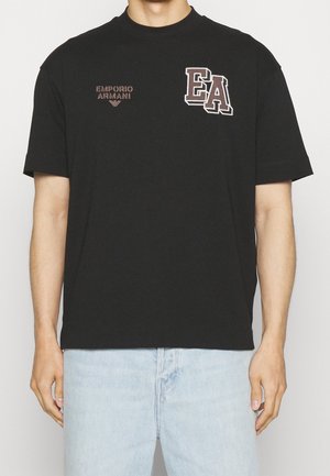 Person trägt ein schwarzes T-Shirt mit kurzen Ärmeln, auf der Brust sind die Aufschriften "EMPORIO ARMANI" und große "EA"-Logos zu sehen, kombiniert mit hellblauen Jeans.