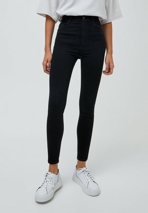 Person trägt hoch taillierte schwarze Skinny-Jeans, weißes oversized T-Shirt, hineingesteckt, und weiße Plateau-Sneaker, steht vor einem einfarbigen Hintergrund.