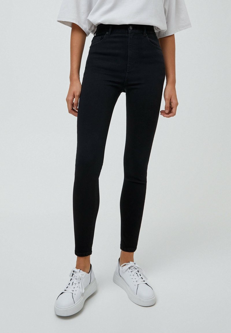 Personne portant un jean skinny noir taille haute, un t-shirt blanc oversize rentré et des baskets blanches à plateforme, debout devant un fond uni.