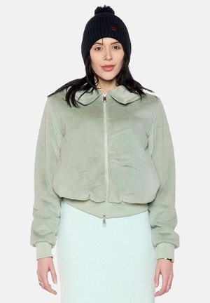 Femme portant une veste pelucheuse vert clair à fermeture éclair et un bonnet en tricot noir avec un pompon, debout devant un fond uni.