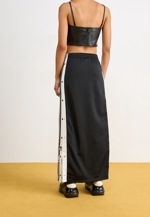 Personne portant un haut court en cuir noir et une longue jupe noire avec des bandes latérales à boutons-pression blancs, ainsi que des chaussures plateformes noires.