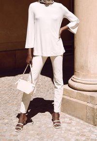 Biała bluzka z długimi rękawami w kształcie dzwonu, zestawiona z białymi dopasowanymi spodniami. Białe sandały na paski i mała torebka dopełniają całość. W tle kamienna kolumna.