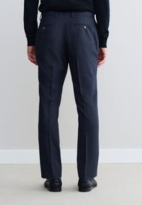 Nohavice po meri, mornarsko modre barve z nežno teksturo, z dvema zadnjima žepoma z gumbi in ravnim krojem. Nošene s črnimi usnjenimi čevlji.