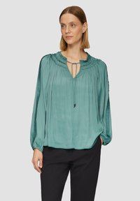 Türkise Bluse mit gefaltetem Detail, weiten Ärmeln und Bindegurtverschluss. Weicher, glatter Stoff, lockere Passform. Kombiniert mit schwarzen Hosen.
