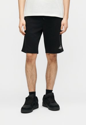 Uomo con pantaloncini neri con un piccolo logo bianco sulla gamba sinistra, calzini neri e scarpe da ginnastica nere, in piedi davanti a uno sfondo semplice.