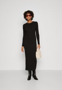 Black long-sleeve dress with a fitted silhouette, paired with knee-high black boots and a small, textured cream handbag.
