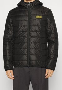 Černá péřová bunda s kapucí, prošívaný design a zipové zapínání. Na hrudi se nachází žluté logo Barbour International. Hladká textura.