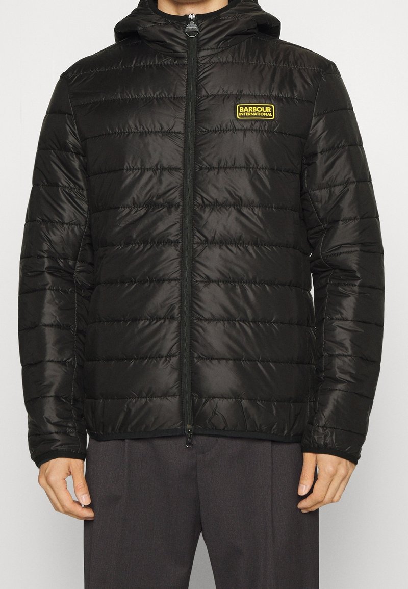 Černá péřová bunda s kapucí, prošívaný design a zipové zapínání. Na hrudi se nachází žluté logo Barbour International. Hladká textura.