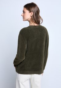 Maglione verde oliva, morbido e con vestibilità rilassata, colletto rotondo e maniche lunghe, realizzato in tessuto soffice e texturizzato. Indossati pantaloni bianchi.