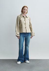 Γυναίκα με κοντά κόκκινα μαλλιά, που φοράει μπεζ σακάκι με κουμπιά, ροζ πουκάμισο με γιακά, καμπάνα μπλε τζιν και λευκά αθλητικά παπούτσια, στέκεται μπροστά σε απλό φόντο.