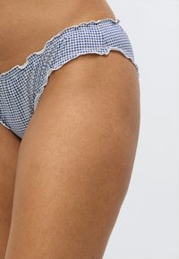 Μπλε και λευκό μπικίνι γκινιάμ με βολάν στα άκρα, που διαθέτει υφασμάτινη υφή και χαμηλή μέση. Το φόντο είναι καθαρό λευκό.