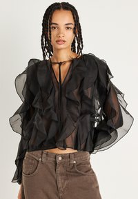 Blusa nera semi-trasparente con strati a volant, maniche lunghe a sbuffo e colletto con fiocco. Abbinata a pantaloni marroni a vita alta.