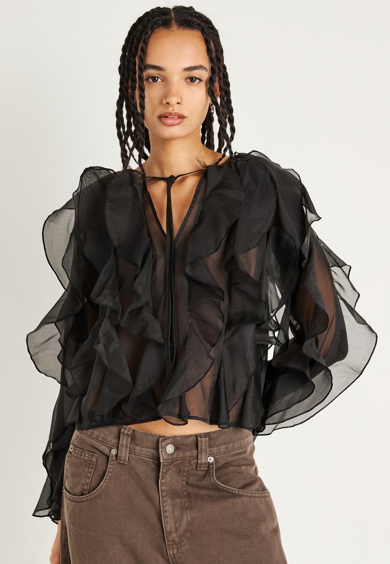 Blusa nera semi-trasparente con strati a volant, maniche lunghe a sbuffo e colletto con fiocco. Abbinata a pantaloni marroni a vita alta.