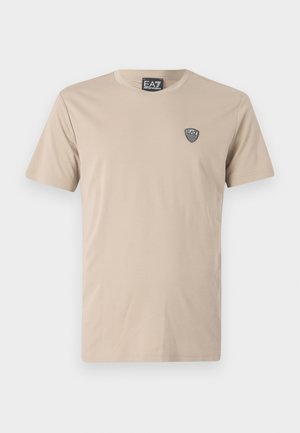 T-shirt en coton beige avec un col rond et des manches courtes, présentant un petit patch logo noir EA7 sur la poitrine gauche. Texture lisse.