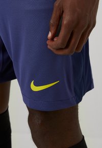 Nike paarse sportshorts van textuurstof, met een geel logo aan de linksonderzijde, en gedeeltelijk zichtbare zwarte kniekousen.
