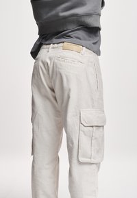 Vue de dos d'une personne portant un pantalon cargo en velours côtelé crème avec poche à rabat et un sweat-shirt gris, debout devant un fond uni.