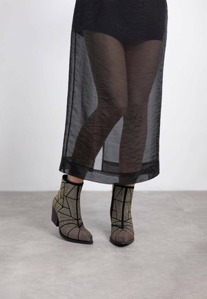 Stivali neri alla caviglia con design metallico geometrico, dotati di tacco a blocco e punta appuntita, abbinati a una gonna nera trasparente.