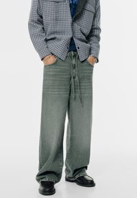 Wijde cargobroek van vervaagde groene denim met een trekkoord in de taille en zijkanten zakken, gecombineerd met een geruit grijs flanellen overhemd en zwarte schoenen.