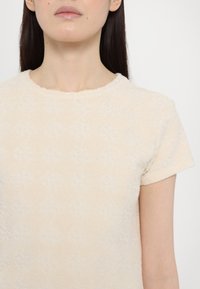 Light beige short-sleeve top in textured fabric, featuring a subtle diamond pattern and a round neckline. Smooth surface, soft feel.