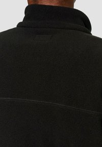 Schwarze Fleecejacke mit hohem Kragen, ausgestattet mit einer strukturierten Oberfläche, Nahtdetails und einem kleinen Logo-Patch am Nacken.