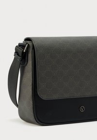 Temno siva in črna torba kurirja z nastavljivim trakom, vzorčastim poklopcem in okroglim logotipom spredaj na enobarvnem ozadju.