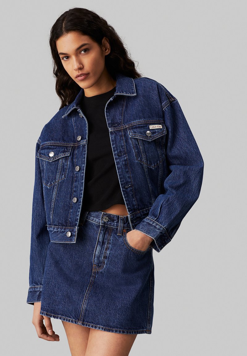 Chaqueta de mezclilla azul oscuro con un corte rectangular y dos bolsillos en el pecho, combinada con una falda mini de mezclilla a juego. Camiseta negra debajo.