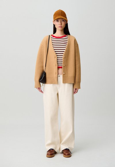 Cárdigan de punto beige, camiseta a rayas roja y blanca, pantalones anchos color crema, zapatos marrones y gorra marrón, con un pequeño bolso negro cruzado.