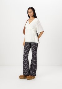 Witte gehaakte wraptop met korte klokmouwen, gecombineerd met zwarte uitlopende broek met witte bloempatronen en bruine schoenen met dikke zolen.