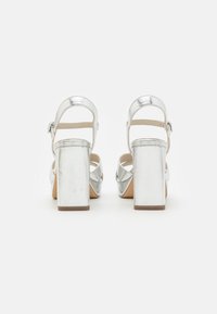Sandalias de tacón alto metálicas plateadas con gruesos tacones cuadrados, una parte trasera abierta y correas en los tobillos. La suela es de color beige, contrastando con el material superior.