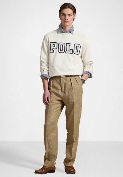 Pulover deschis cu crem cu inscriptionarea "POLO" în litere bleumarin, suprapus peste o cămașă cu guler în carouri albastre și albe. Pantaloni închis cu carouri bej și pantofi maro.