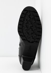 Semelle de chaussure noire avec un motif de bande de roulement profond comprenant des croix et des barres horizontales, légèrement inclinée sur un fond blanc en angle.