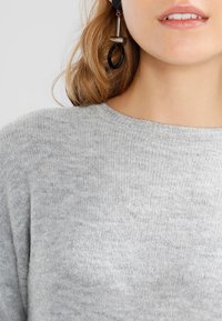 Pull en maille texturée gris clair avec un col rond et un tissu doux. Des boucles d'oreilles pendantes noires et métalliques viennent rehausser la tenue.