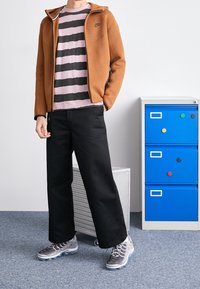 Hoodie zippé marron, t-shirt rose à rayures noires, pantalon noir ample et baskets grises avec des accents métalliques. Armoire bleue en arrière-plan.