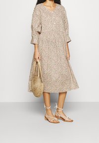 Vestido midi floral con escote en V, mangas tres cuartos y detalle de botones; presenta un fondo crema con flores rosas y verdes. Sandalias tostadas y bolso de paja.