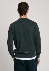 Sudadera verde oscura de tela suave, con cuello redondo, mangas largas y un detalle de logo en el dobladillo trasero. Corte casual y acabado texturizado.