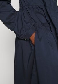 Mörkblå, lättviktsklänning med samlad midja. Har långa ärmar och sidofickor. Slät textur, utan mönster eller dekorationer.