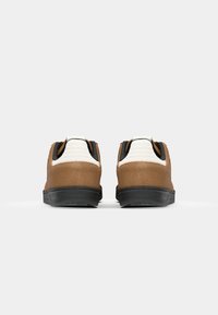 Zapatos de ante marrón con acentos de textil blanco, suelas de goma negra y un diseño curvado. Cuentan con una lengüeta en el talón para facilitar el uso.