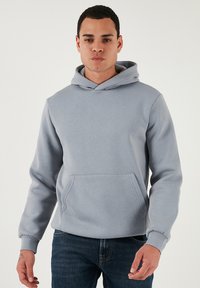 Szürke kapucnis pulcsi kenguruzsákkal, puha anyagból készült. Húzózsinórral ellátott kapucni és bordázott mandzsetták jellemzik. Kék farmerrel viselve.