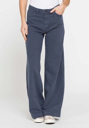 Persona che indossa pantaloni a palazzo blu navy e sneaker bianche, con una mano in tasca, in piedi davanti a uno sfondo chiaro e semplice.