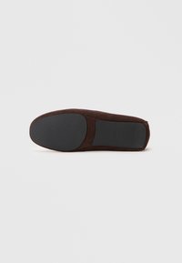 Suela de pantufla de ante marrón con parches de goma negra en el talón y el dedo del pie, exhibida plana sobre un fondo blanco.