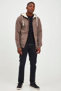 Sudadera con cremallera marrón y forro interior crema, que incluye un bolsillo frontal y cordones, combinada con jeans oscuros y zapatillas negras.