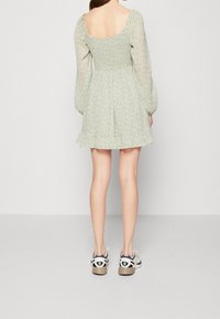 Kvinna som står med ryggen mot oss, iklädd en ljusgrön blommig klänning med puffärmar och vita sneakers.