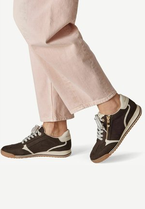 Person wearing beige pants and black and white sneakers with side zipper and white laces, standing on white background.