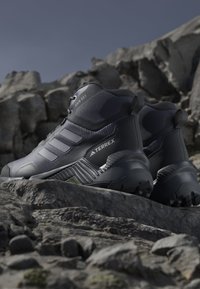 Black and grey Adidas Terrex hiking boots with Rain.RDY waterproofing, placed on rocky terrain under clear sky.