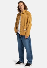 Lichtbruine knoop-up jas met een borstzak, gecombineerd met losse, vervaagde blauwe jeans en zwarte skate schoenen. Effen wit T-shirt eronder.